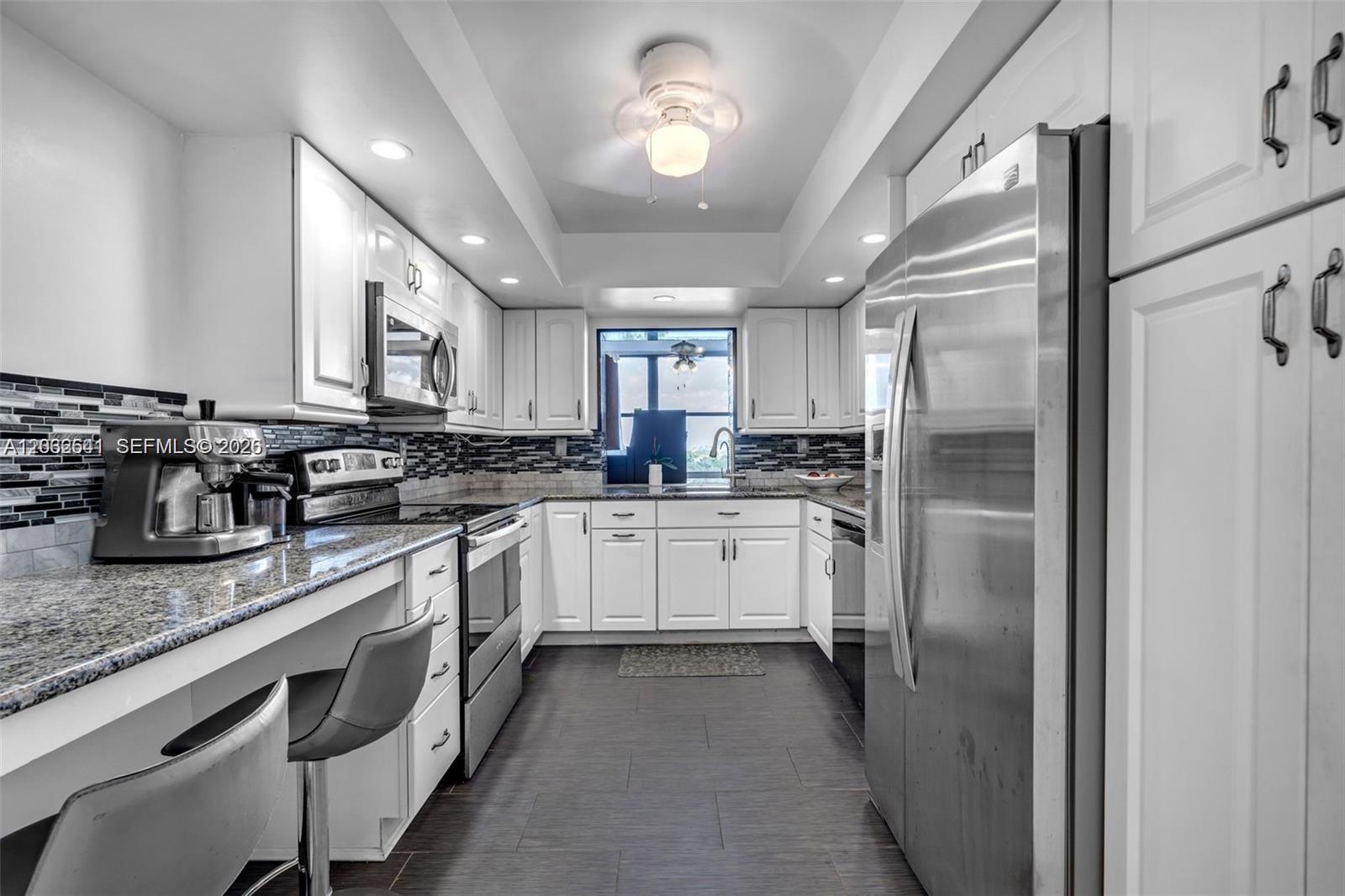 A galley kitchen features white cabinetry, stainless steel appliances, granite countertops, and dark tile flooring — 1000 Parkview Dr # 715, Hallandale Beach FL 33009, HALLANDALE BEACH, FL