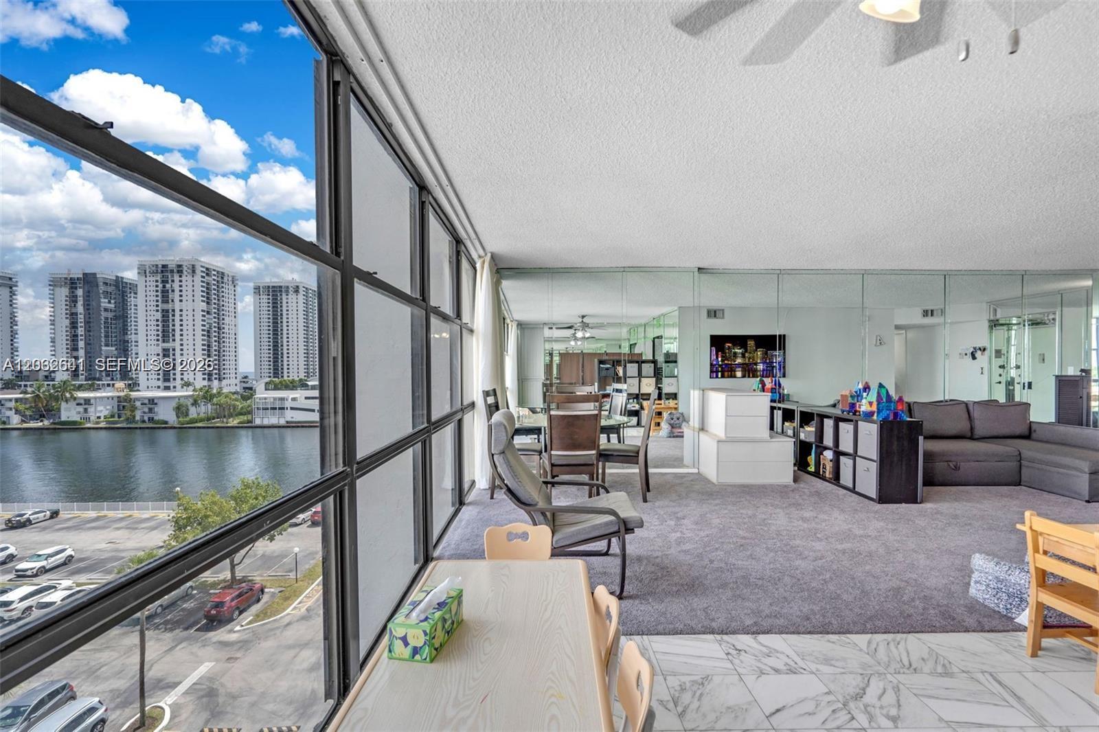 A living area with gray carpet, mirrored walls, and floor-to-ceiling windows overlooking a waterfront view — 1000 Parkview Dr # 715, Hallandale Beach FL 33009, HALLANDALE BEACH, FL