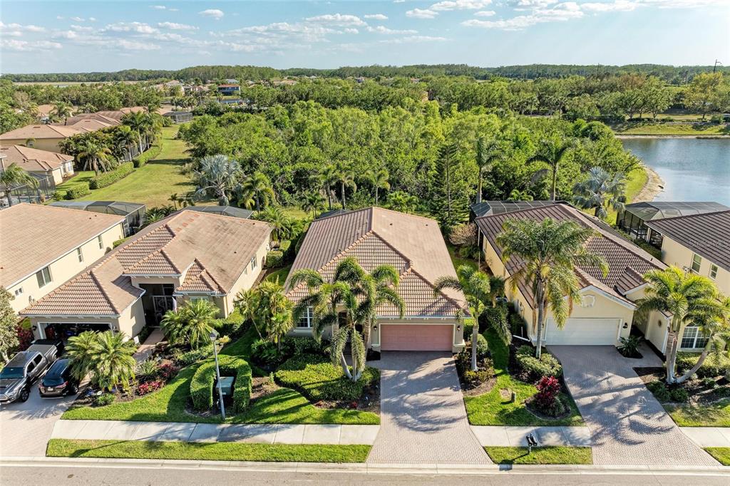 An aerial view shows three suburban houses with tiled roofs, palm trees, and a nearby lake — 135 RIVER ENCLAVE COURT, BRADENTON, FL
