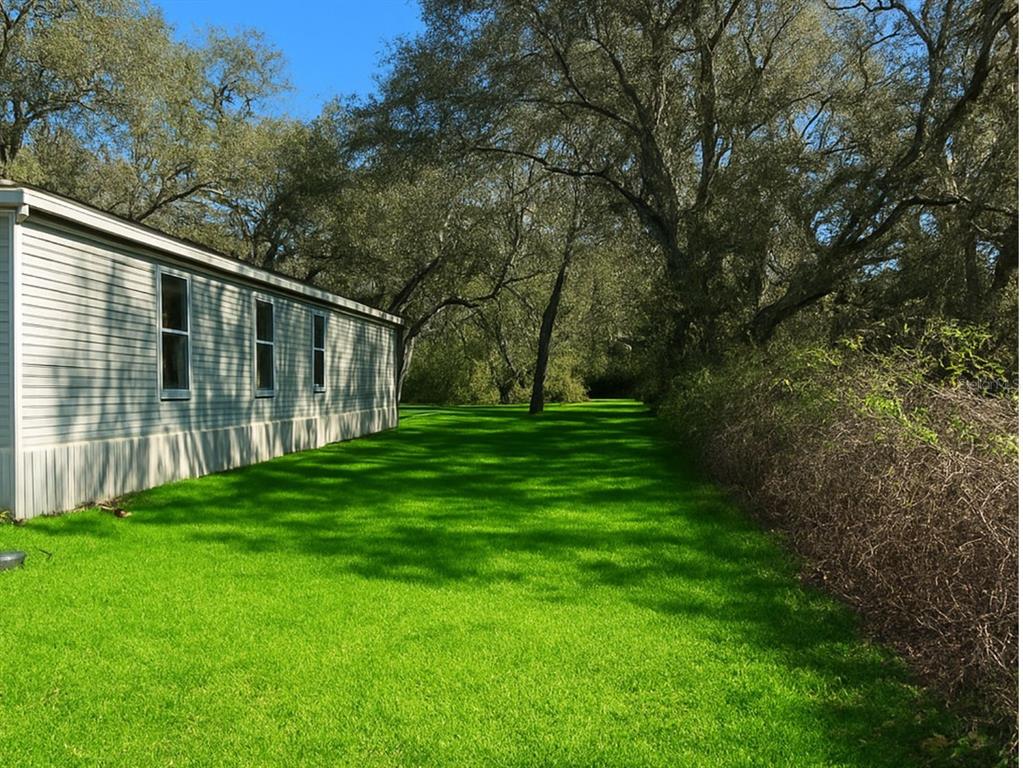 A side view of a light-colored mobile home beside a grassy yard and mature trees — 14640 NE 149TH LANE, FORT MC COY, FL