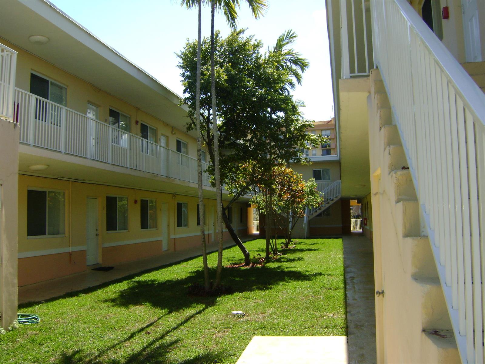 A grassy courtyard sits between two levels of apartment buildings with white railings and stairs — NORTH MIAMI BEACH, FL