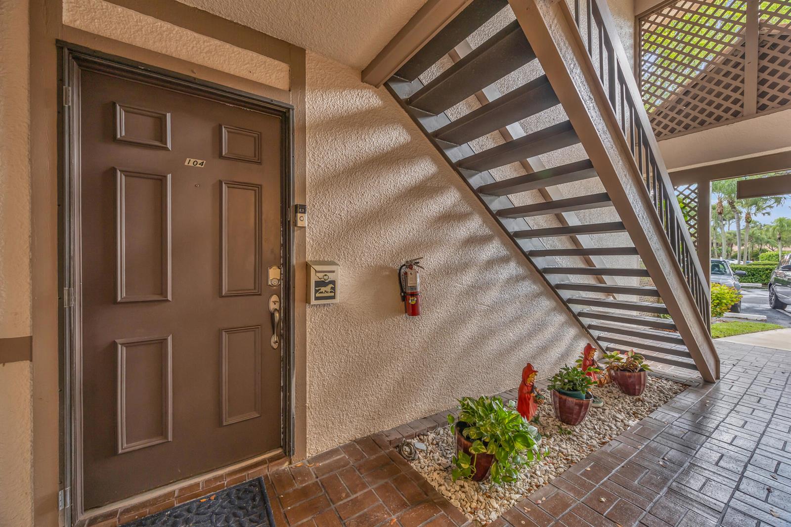 An exterior view of a brown apartment door labeled 104 with stairs and potted plants — 1105 Duncan Circle # 104, Palm Beach Gardens FL 33418, PALM BEACH GARDENS, FL