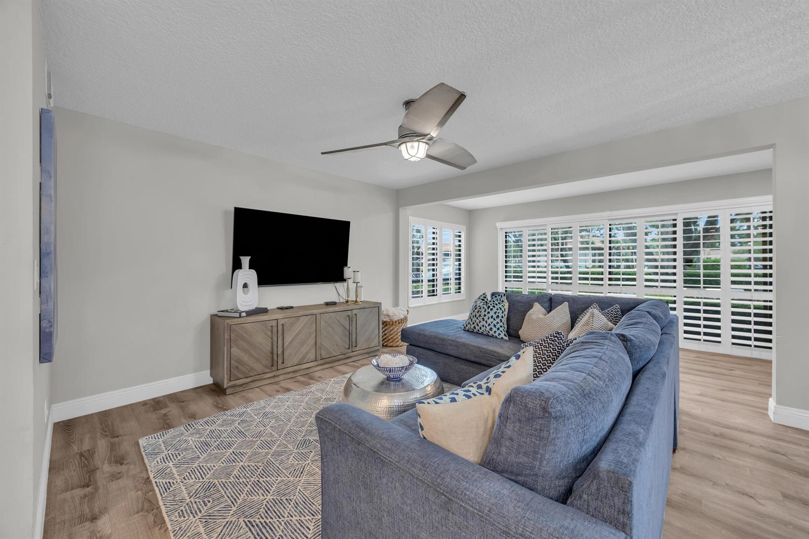 A living room features a blue sectional sofa, a television, and plantation shutters — 1105 Duncan Circle # 104, Palm Beach Gardens FL 33418, PALM BEACH GARDENS, FL