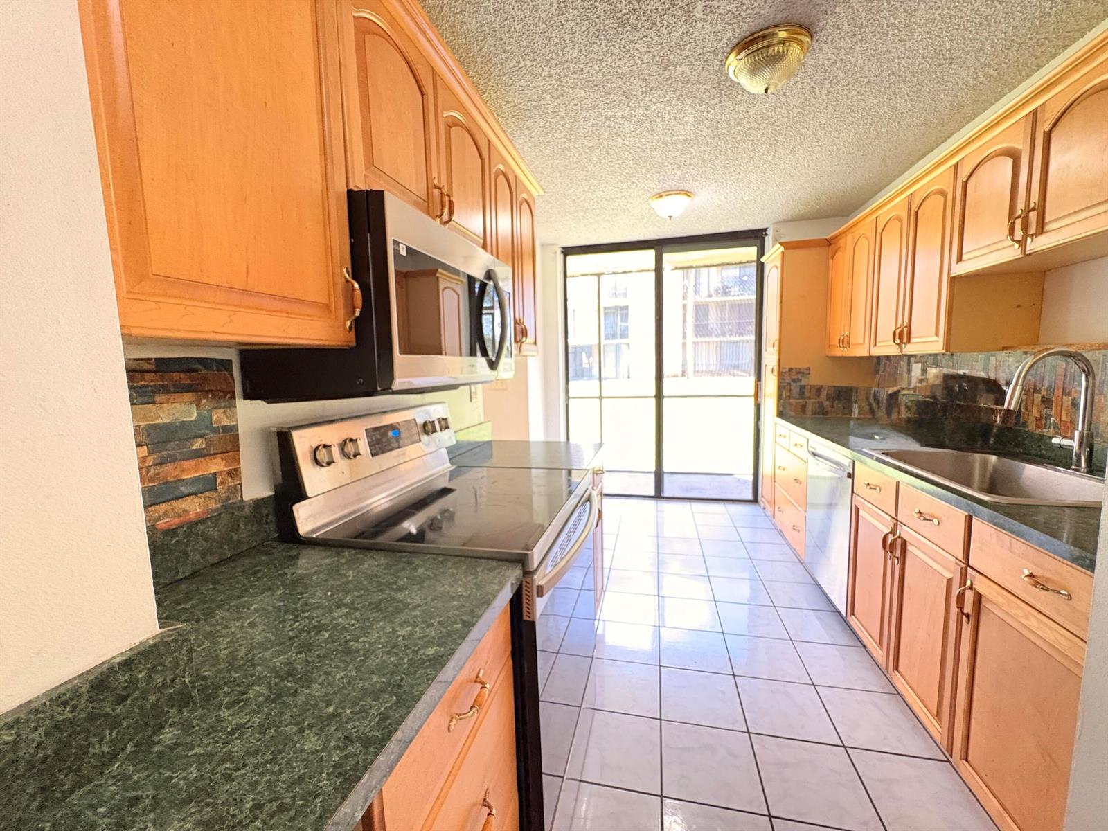 A galley kitchen features light wood cabinets, green laminate countertops, and a sliding glass door — 3590 Blue Lake Drive # 104, Pompano Beach FL 33064, POMPANO BEACH, FL