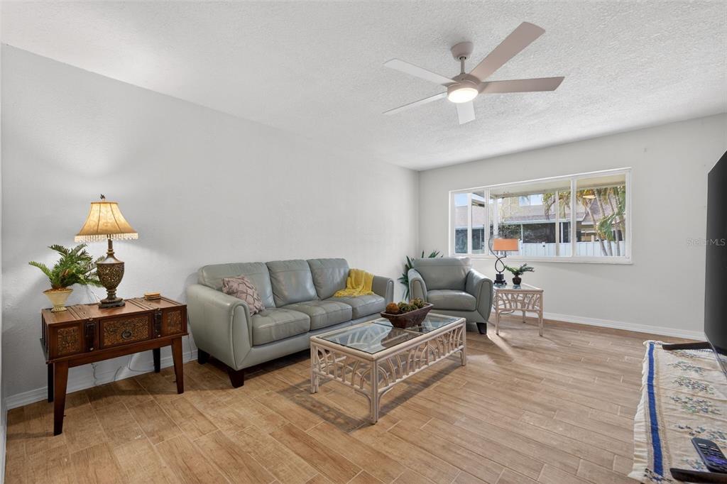 A living room features a gray sofa, matching armchair, glass-topped coffee table, and wood-look tile flooring — 6490 CAPE HATTERAS WAY NE UNIT 2, ST PETERSBURG, FL