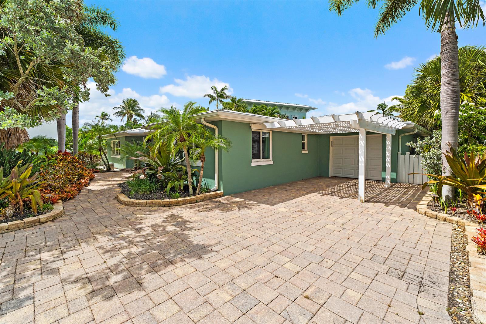 A green house with a white pergola over a paved driveway and tropical landscaping — 440 Jupiter Lane, Juno Beach FL 33408, JUNO BEACH, FL