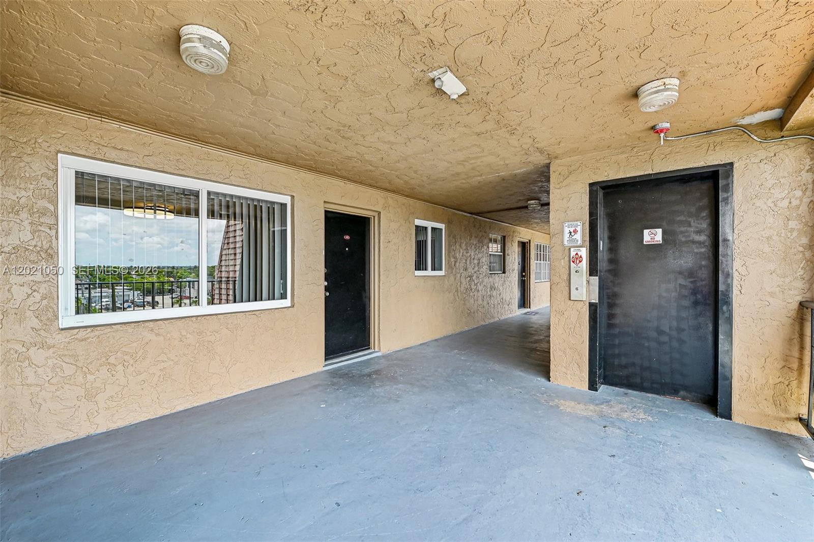 An outdoor apartment hallway features textured tan walls, a gray floor, and black doors — 6575 W 4th Ave # 507, Hialeah FL 33012, HIALEAH, FL