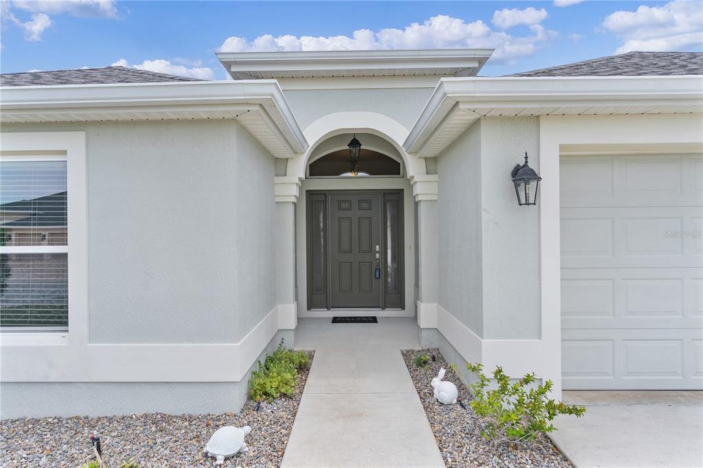 The exterior of a house features a gray front door, a concrete walkway, and landscaping — 5833 ABNEY AVENUE, THE VILLAGES, FL