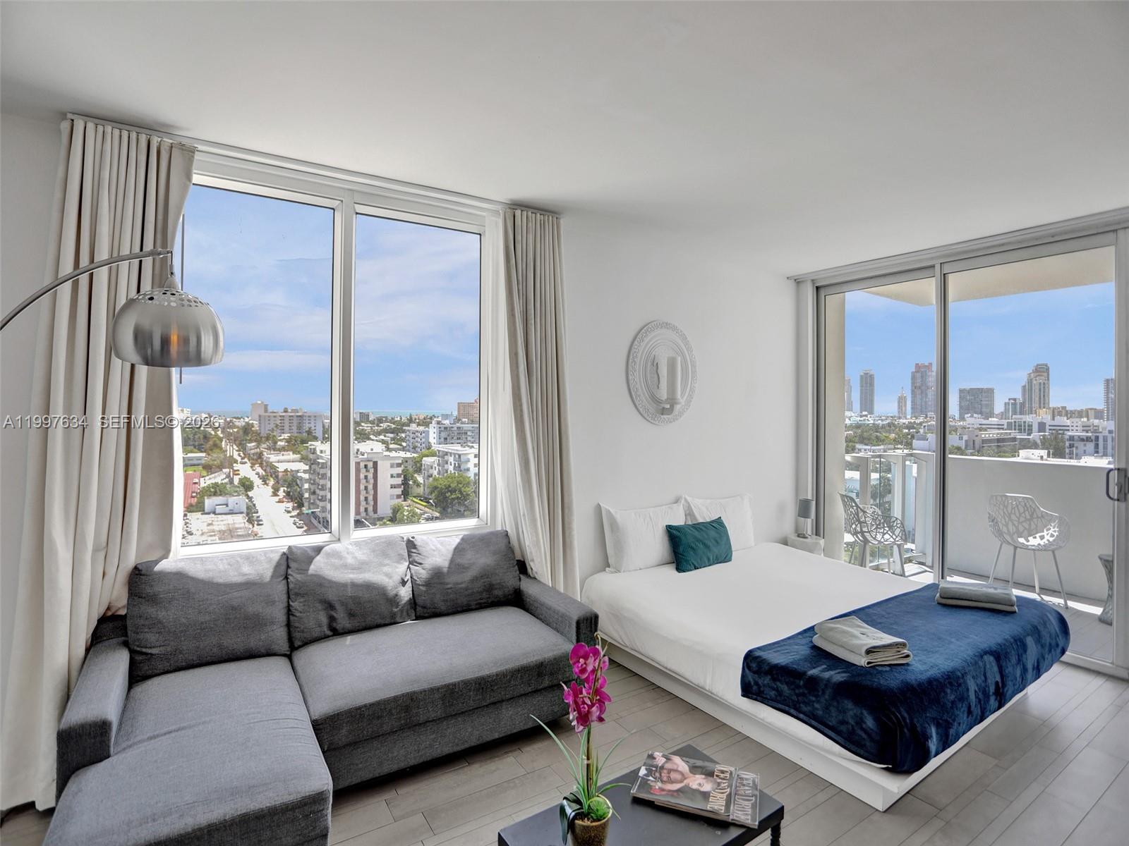A studio apartment features a gray sectional sofa, a bed, and floor-to-ceiling windows with city views — 1100 West Ave # 1101, Miami Beach FL 33139, MIAMI BEACH, FL