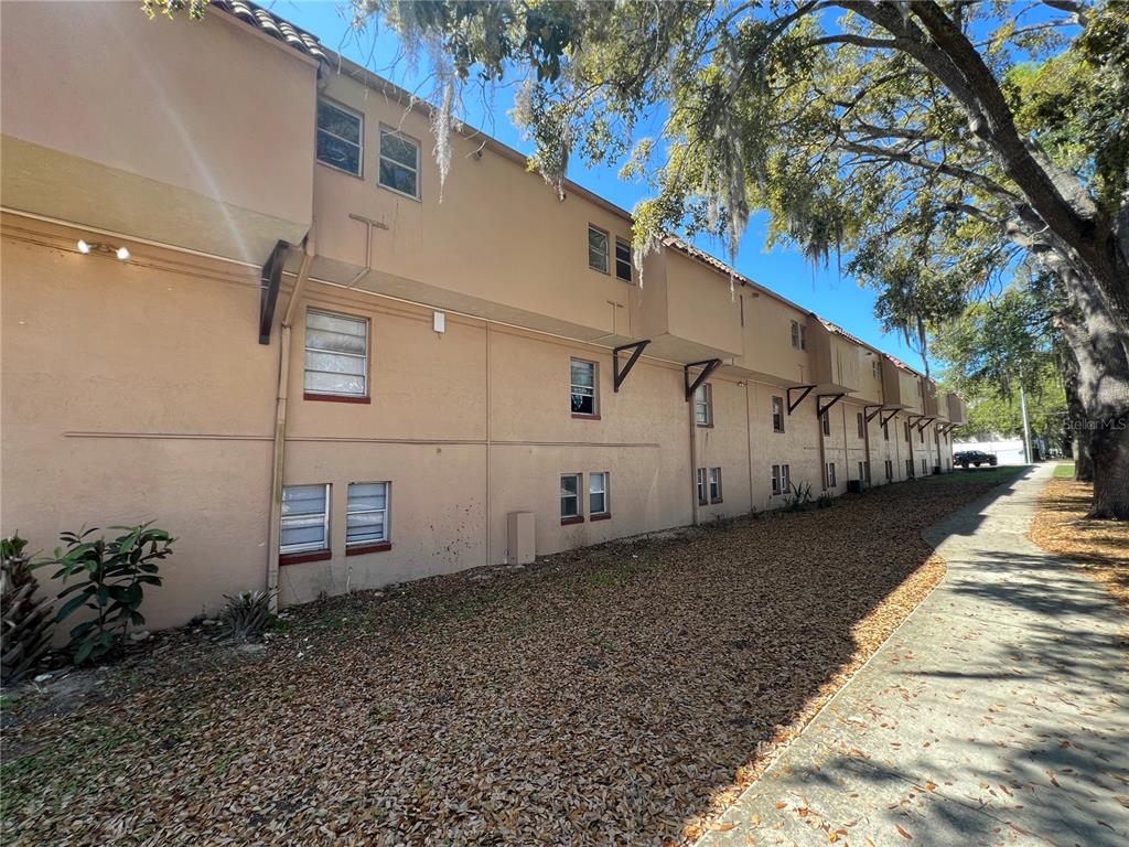 A side view of a multi-story tan stucco apartment building with a sidewalk and trees — 914 SW 8TH AVENUE UNIT 44, GAINESVILLE, FL