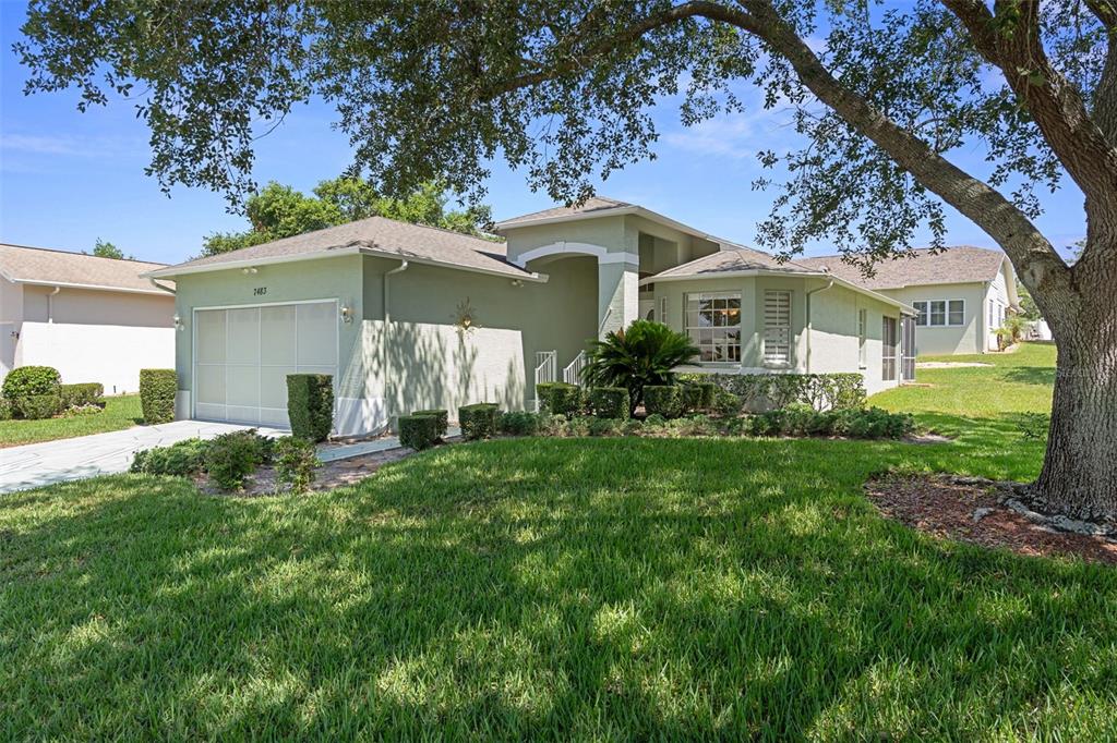 A light green single-story house with a two-car garage and a large front yard tree — 7483 BRIDGEWATER LANE, SPRING HILL, FL