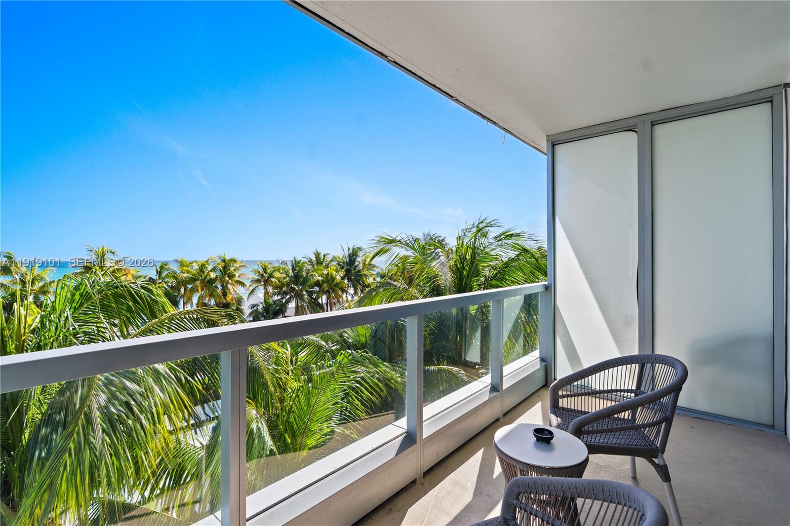 A balcony features a glass railing, two woven chairs, a small table, and palm trees — 4391 Collins Ave # 413, Miami Beach FL 33140, MIAMI BEACH, FL