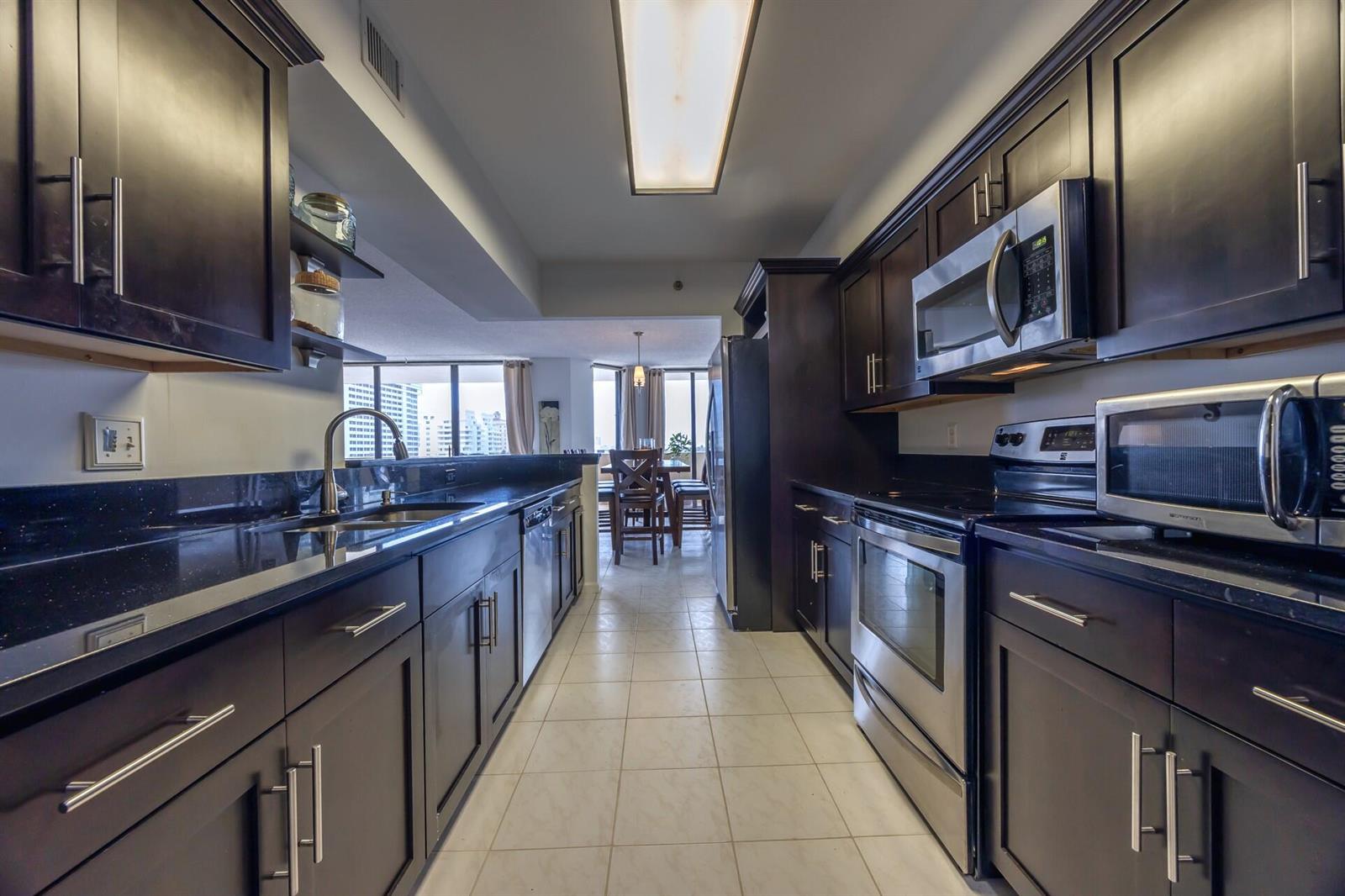 A galley kitchen features dark wood cabinets, black countertops, and stainless steel appliances — 1401 S Ocean Boulevard # 5030, Boca Raton FL 33432, BOCA RATON, FL