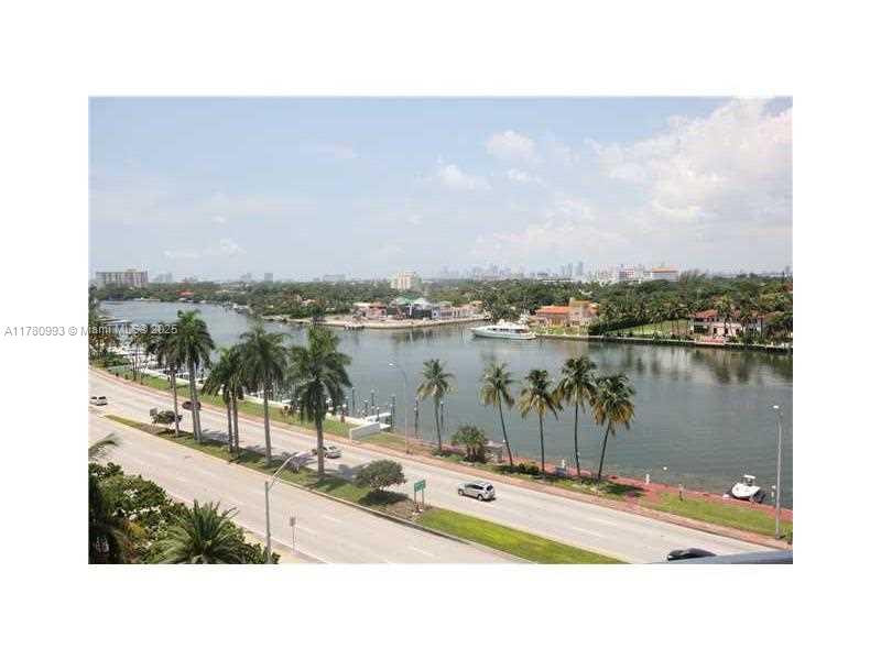 An elevated view of a waterway, a coastal road, palm trees, and distant city buildings — 5151 COLLINS AV # 924, Miami Beach FL 33140, MIAMI BEACH, FL