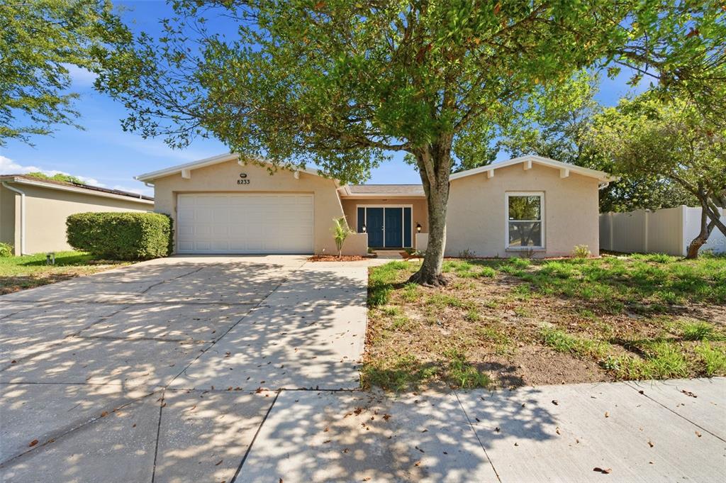 A single-story house with a beige stucco exterior, a two-car garage, and a large tree — 8233 REDFIELD DRIVE, PORT RICHEY, FL