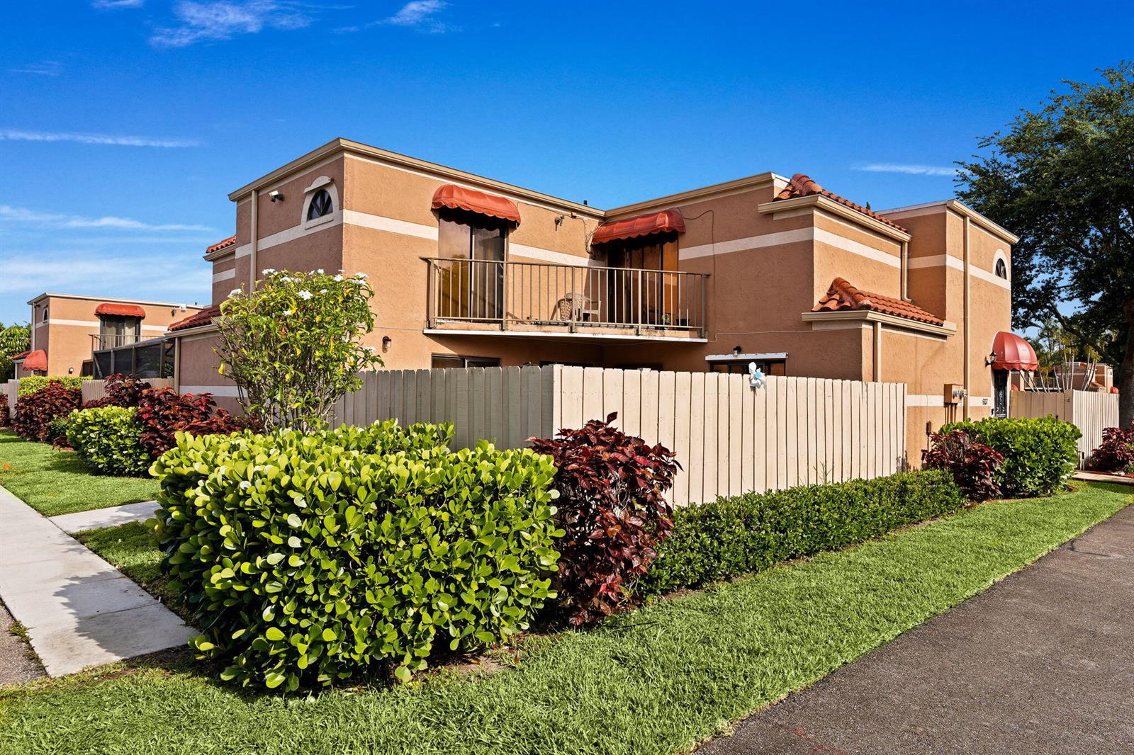 An exterior view of a two-story tan stucco townhouse with a balcony and wooden fence — 6139 Seven Springs Boulevard, FL 33463, FL
