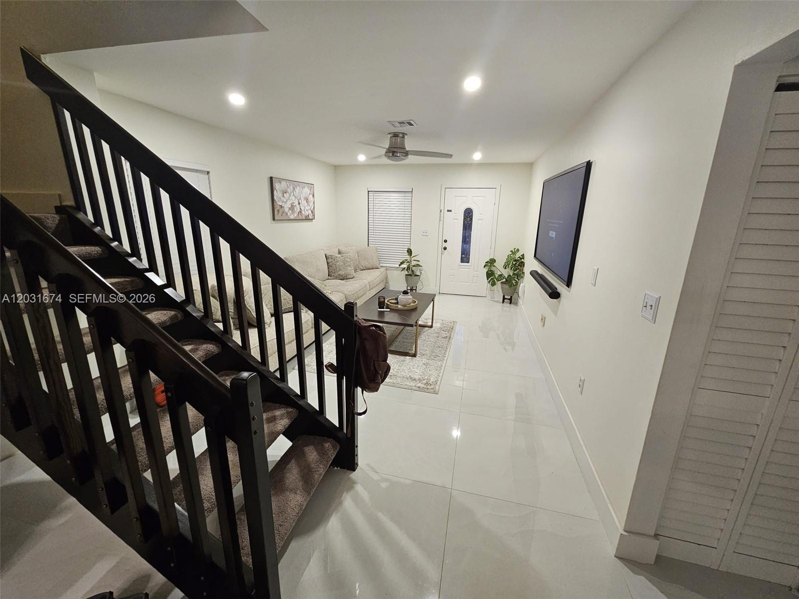 A living room features a dark wooden staircase, white tile floors, and a wall-mounted television — MIRAMAR, FL