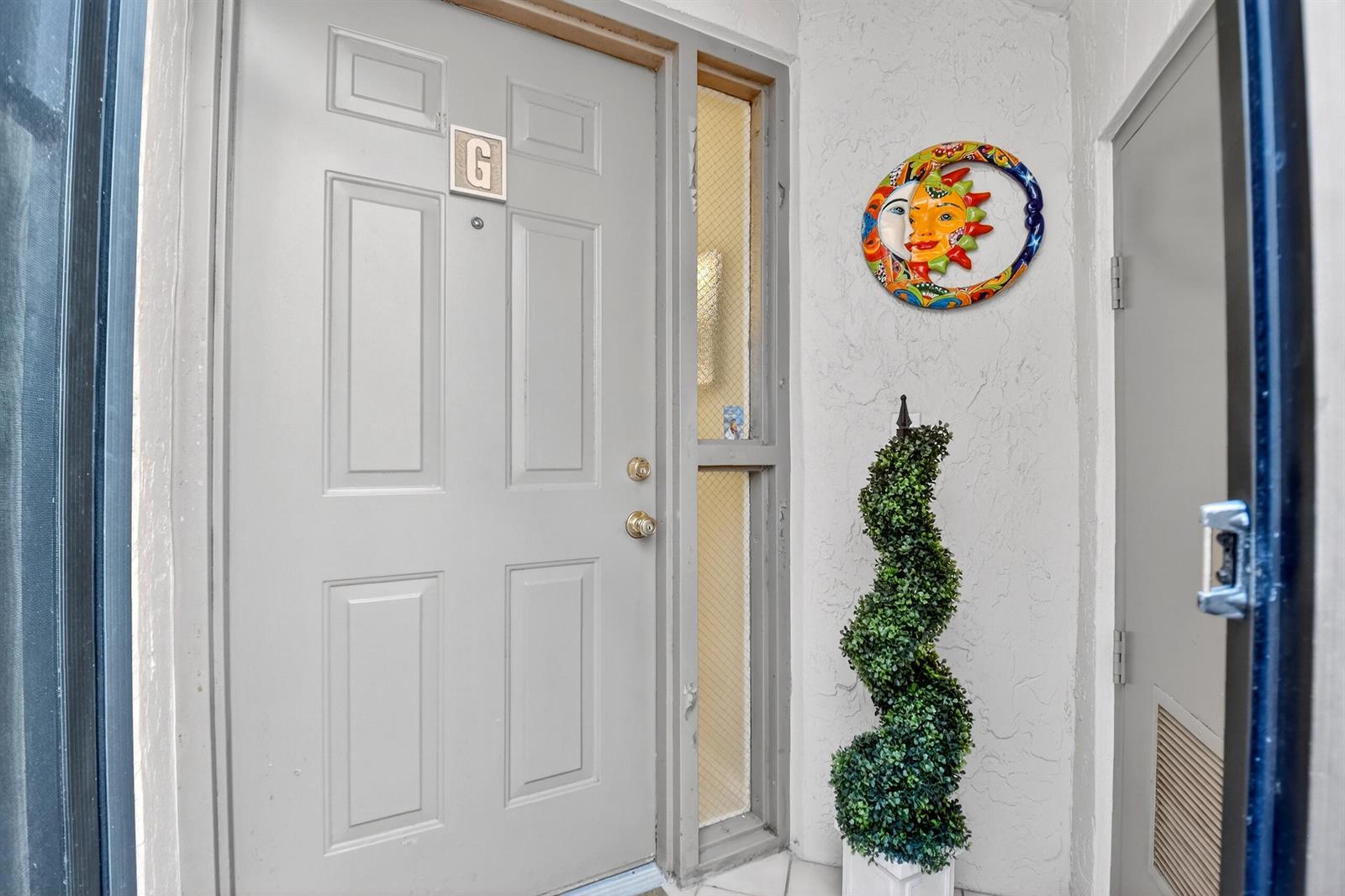 A gray front door with a side window and a decorative sun wall hanging — 5188 Europa Drive # G, Boynton Beach FL 33437, BOYNTON BEACH, FL