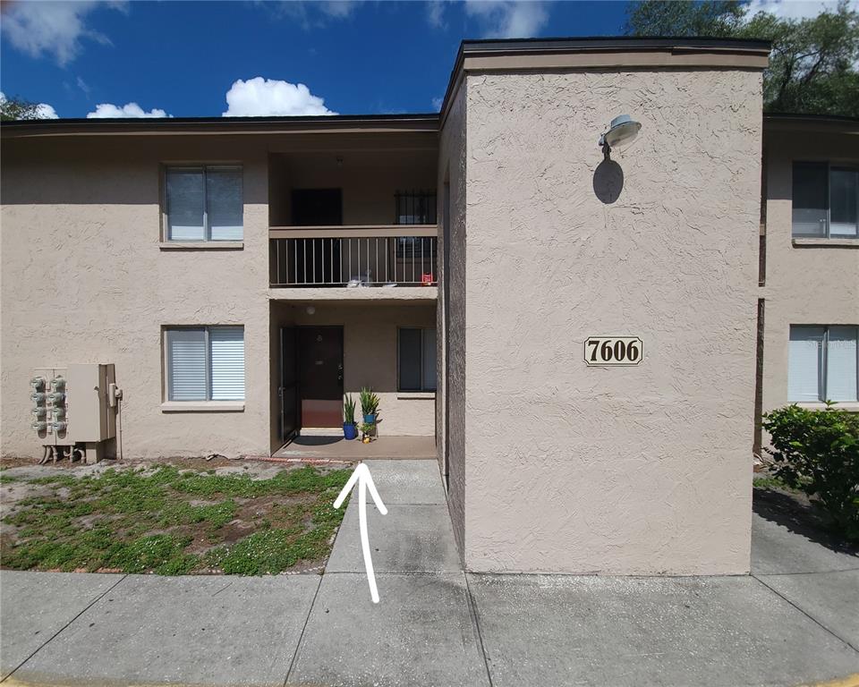 A two-story tan stucco apartment building exterior with a ground-level entrance and a balcony above — 7606 ABBEY LANE UNIT 208, TAMPA, FL