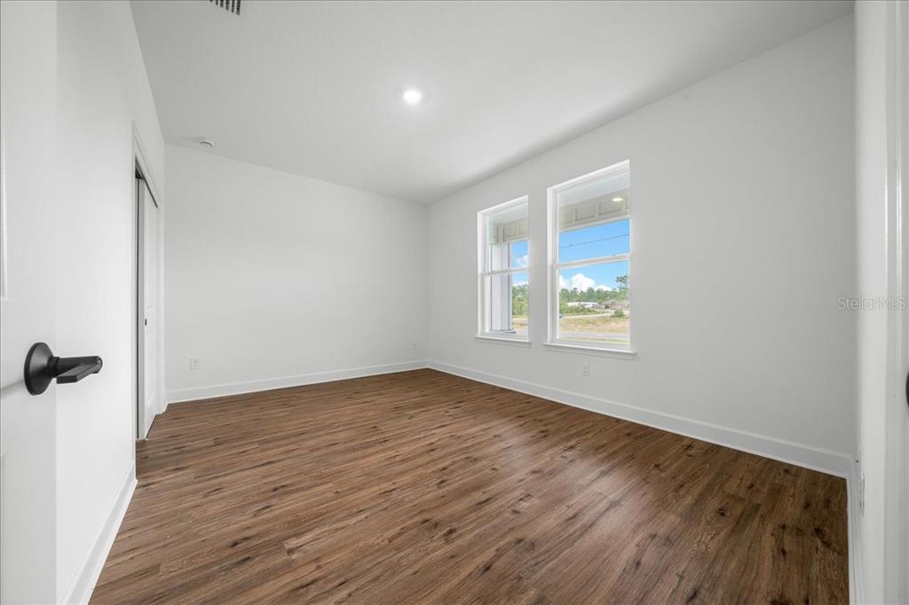 An empty bedroom features white walls, wood-look flooring, and two windows overlooking a construction site — 16213 PENN STATE ROAD, WEEKI WACHEE, FL