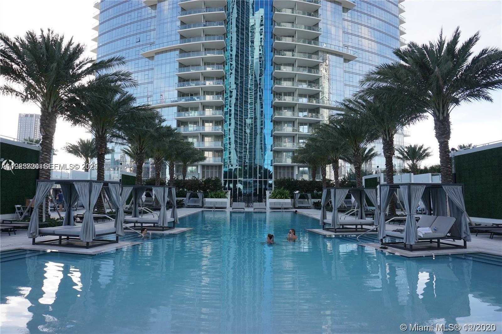 A swimming pool is surrounded by cabanas and palm trees in front of a glass skyscraper — 851 NE 1st Ave # 4911, Miami FL 33132, MIAMI, FL
