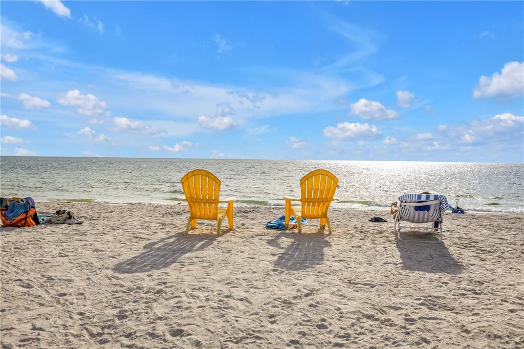 Two yellow Adirondack chairs and a lounge chair sit on a sandy beach by water — 134 86TH TERRACE, TREASURE ISLAND, FL