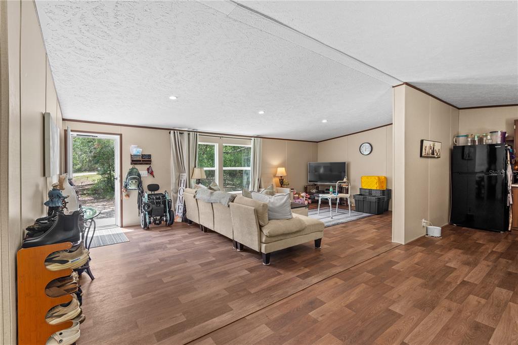 An open-concept living room with wood-look flooring, a beige sectional sofa, and a black refrigerator — 8551 NE 102ND COURT, BRONSON, FL