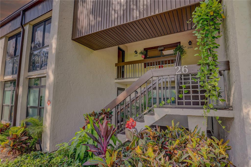 Exterior view of a two-story apartment building with a wooden staircase and lush landscaping — 2400 WINDING CREEK BOULEVARD UNIT 26-203, CLEARWATER, FL