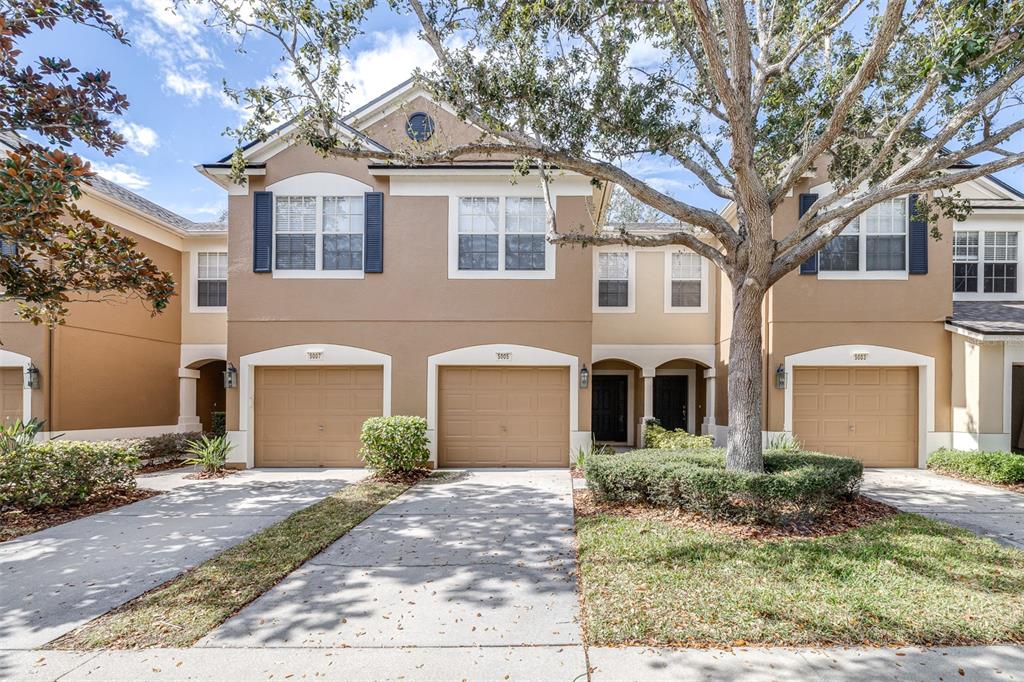 A tan two-story townhouse exterior features three garage doors, shuttered windows, and a large tree — 5005 BARNSTEAD DRIVE, RIVERVIEW, FL