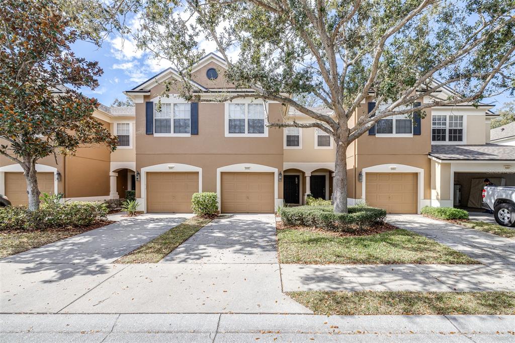 A row of tan townhomes with attached garages and driveways under a large tree — 5005 BARNSTEAD DRIVE, RIVERVIEW, FL