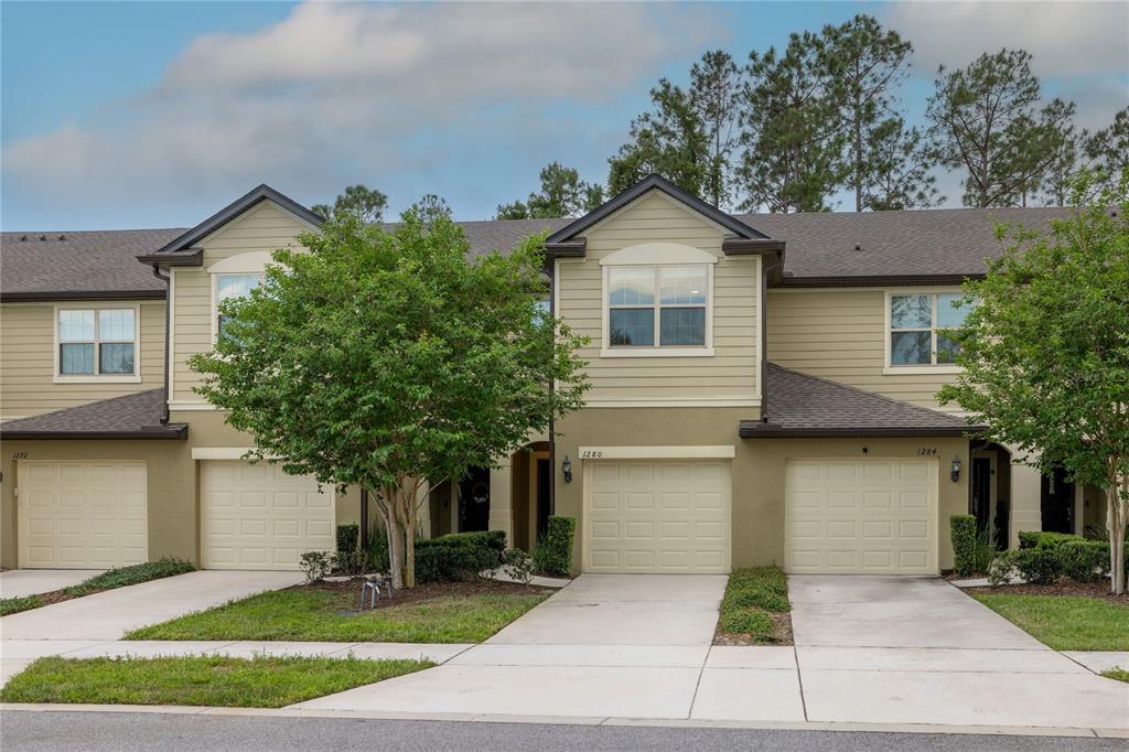 A row of three attached townhomes with beige siding, garages, and front-facing trees — 1280 PAVIA DRIVE, APOPKA, FL