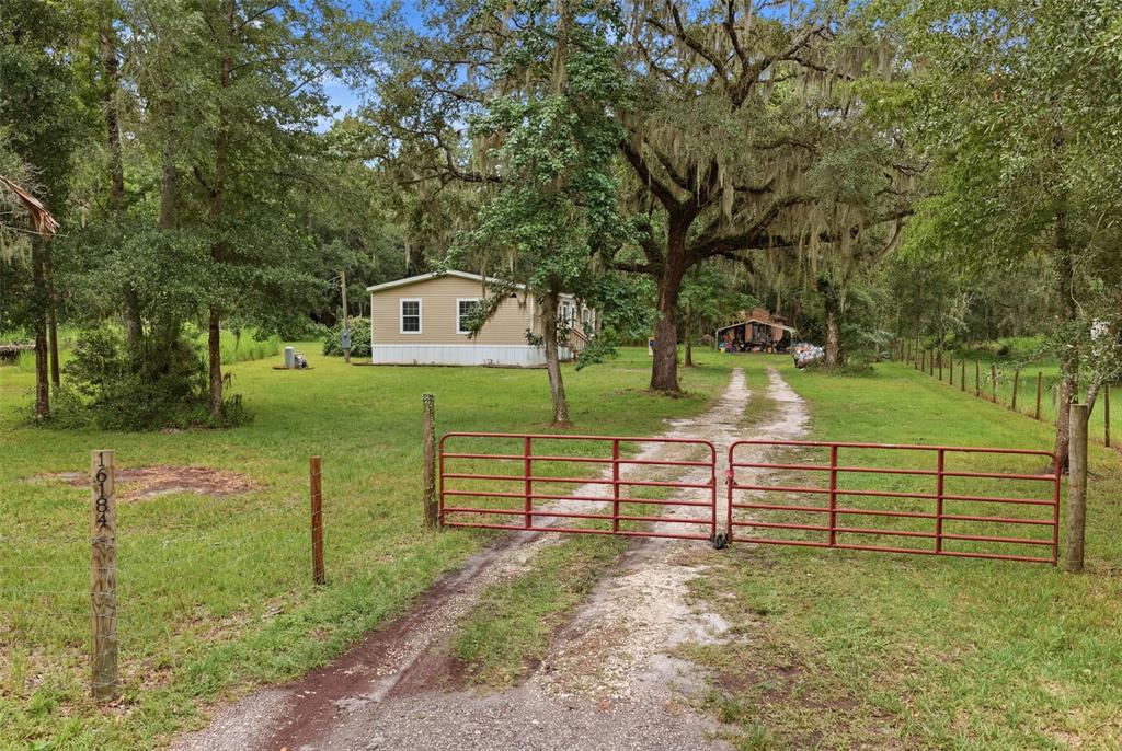 A red metal gate opens to a dirt driveway leading toward a tan mobile home — 16184 PAXFORD LANE, BROOKSVILLE, FL