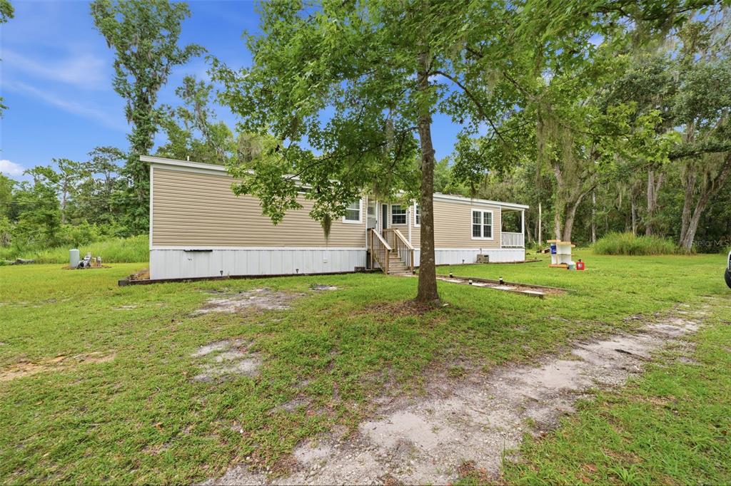 A tan manufactured home with a wooden staircase sits on a grassy lot with trees — 16184 PAXFORD LANE, BROOKSVILLE, FL