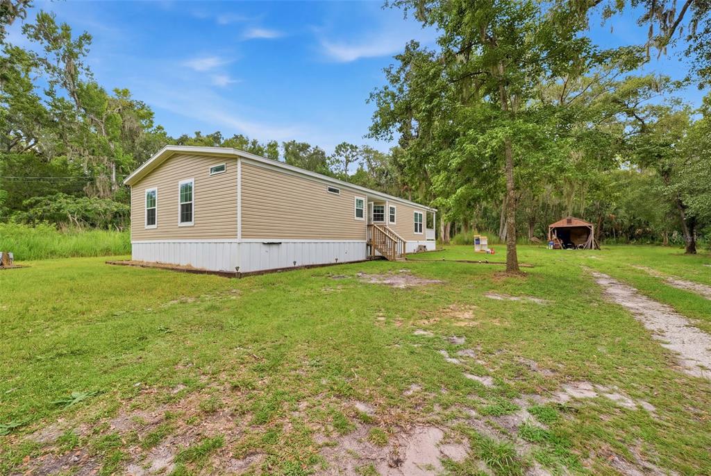 A tan manufactured home with white skirting sits on a grassy lot with trees — 16184 PAXFORD LANE, BROOKSVILLE, FL