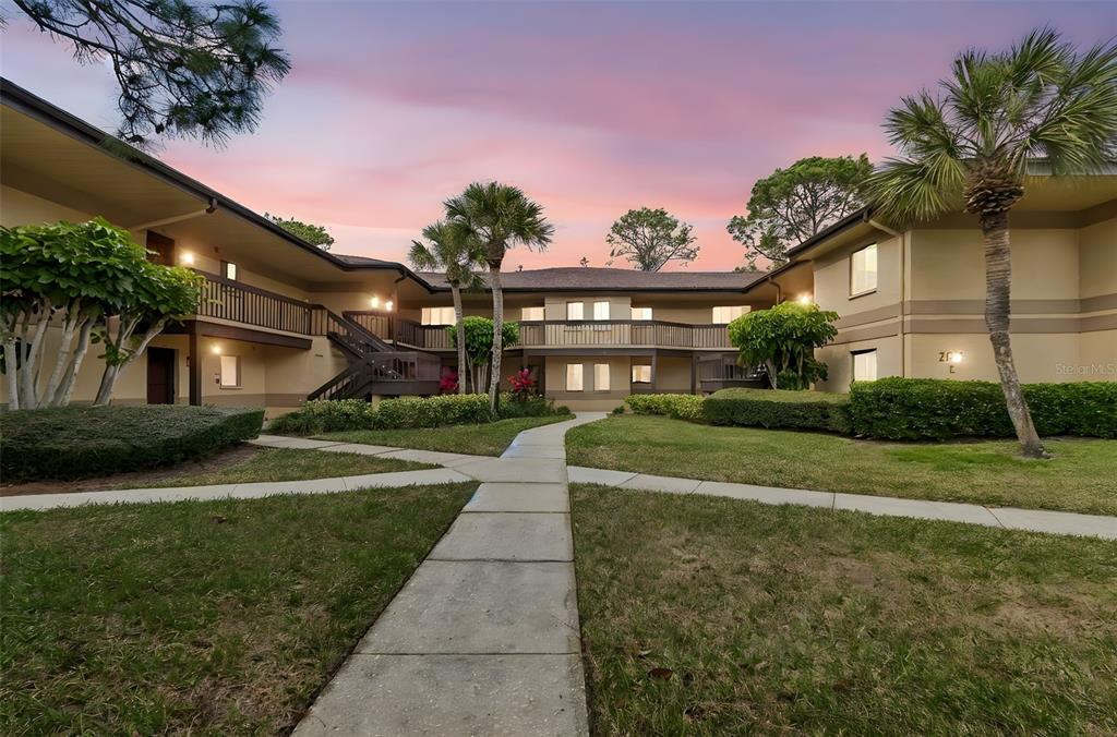 A two-story apartment building exterior with a central walkway, palm trees, and a sunset sky — 2679 SABAL SPRINGS CIRCLE UNIT 102, CLEARWATER, FL