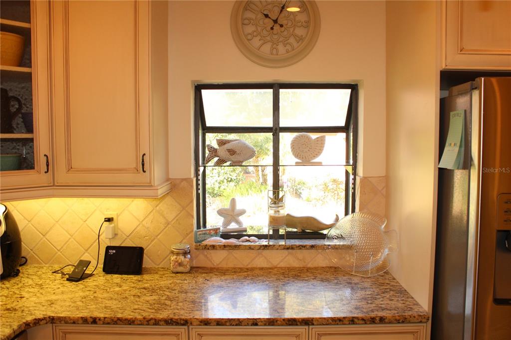 A kitchen features light-colored cabinets, granite countertops, a window with coastal decor, and a clock — 7909 4TH AVENUE W UNIT 12, BRADENTON, FL