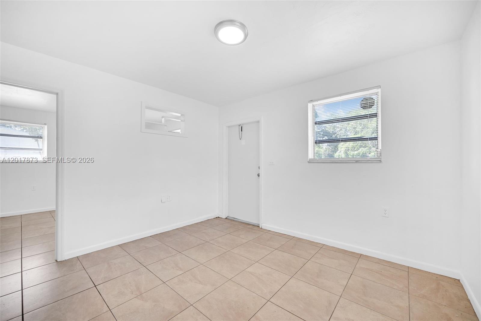 A sparsely furnished room features white walls, tan tile flooring, a window, and a door — 567-3 NW 90th St, El Portal FL 33150, EL PORTAL, FL