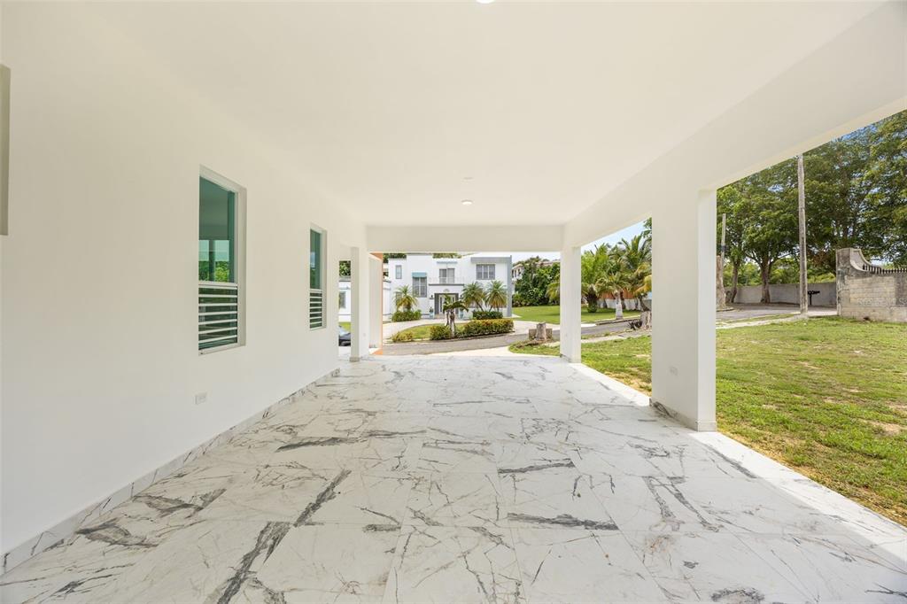 A covered patio with white marble-patterned tile flooring overlooks a grassy yard and neighboring house — solar 2 villa alemeria MARCEL, AGUADILLA, PR