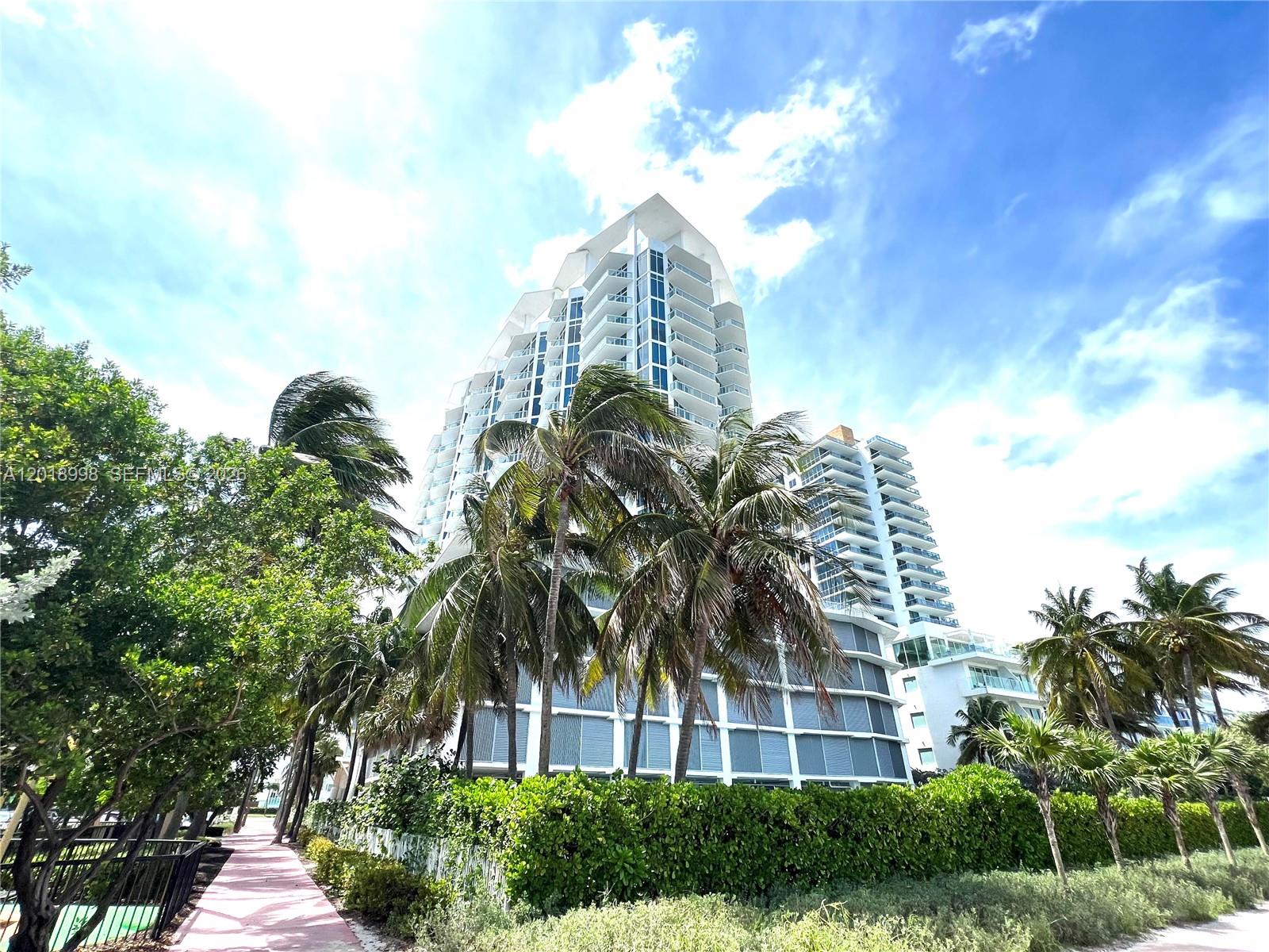A low-angle view of a modern white high-rise condominium building surrounded by palm trees — 6515 Collins Ave # 1801, Miami Beach FL 33141, MIAMI BEACH, FL