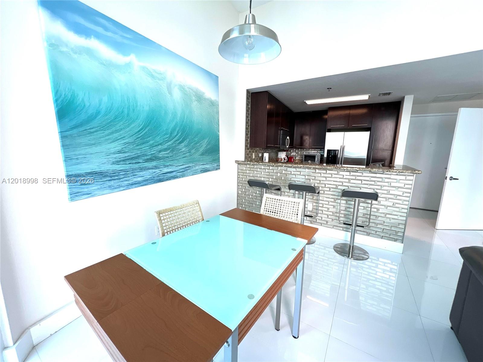 A dining area features a glass-topped table, bar seating, and a large ocean wave painting — 6515 Collins Ave # 1801, Miami Beach FL 33141, MIAMI BEACH, FL