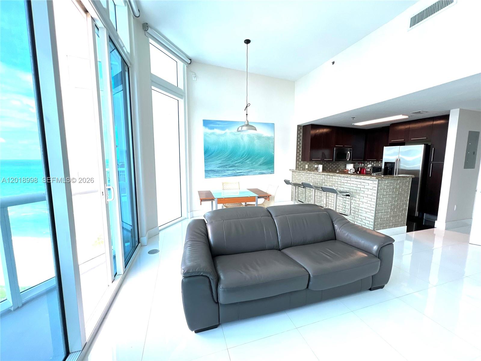 A modern living room and kitchen area features a grey sofa, ocean view, and tile floors — 6515 Collins Ave # 1801, Miami Beach FL 33141, MIAMI BEACH, FL