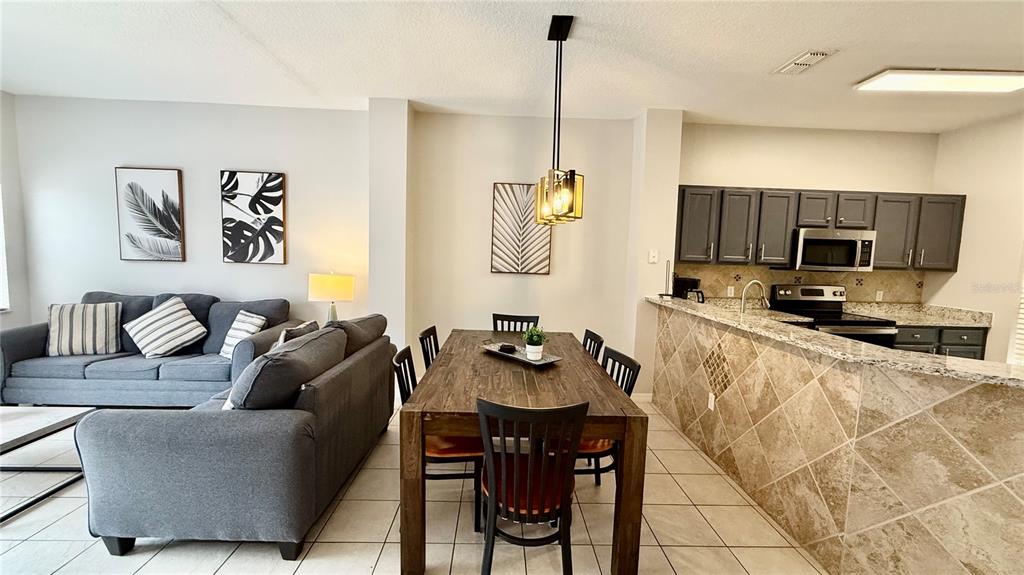 An open-concept living and dining area features a grey sofa, wooden table, and tiled kitchen — 8517 BAY LILLY LOOP, KISSIMMEE, FL