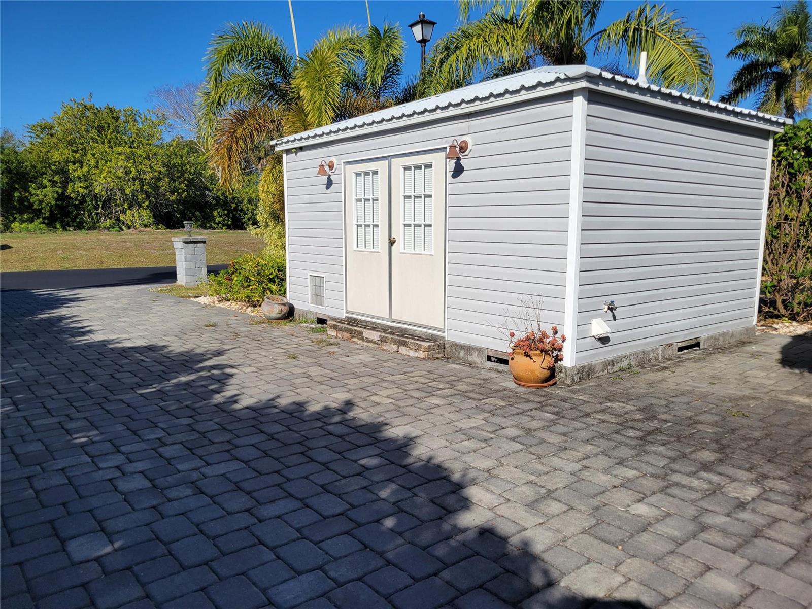 A gray vinyl-sided shed with double doors sits on a paved patio near palm trees — 3528 SW 7th Manor, Okeechobee FL 34974, OKEECHOBEE, FL