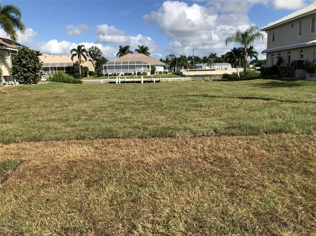 A vacant grassy lot sits in front of a canal with houses in the background — 3754 SPOONBILL COURT, PUNTA GORDA, FL