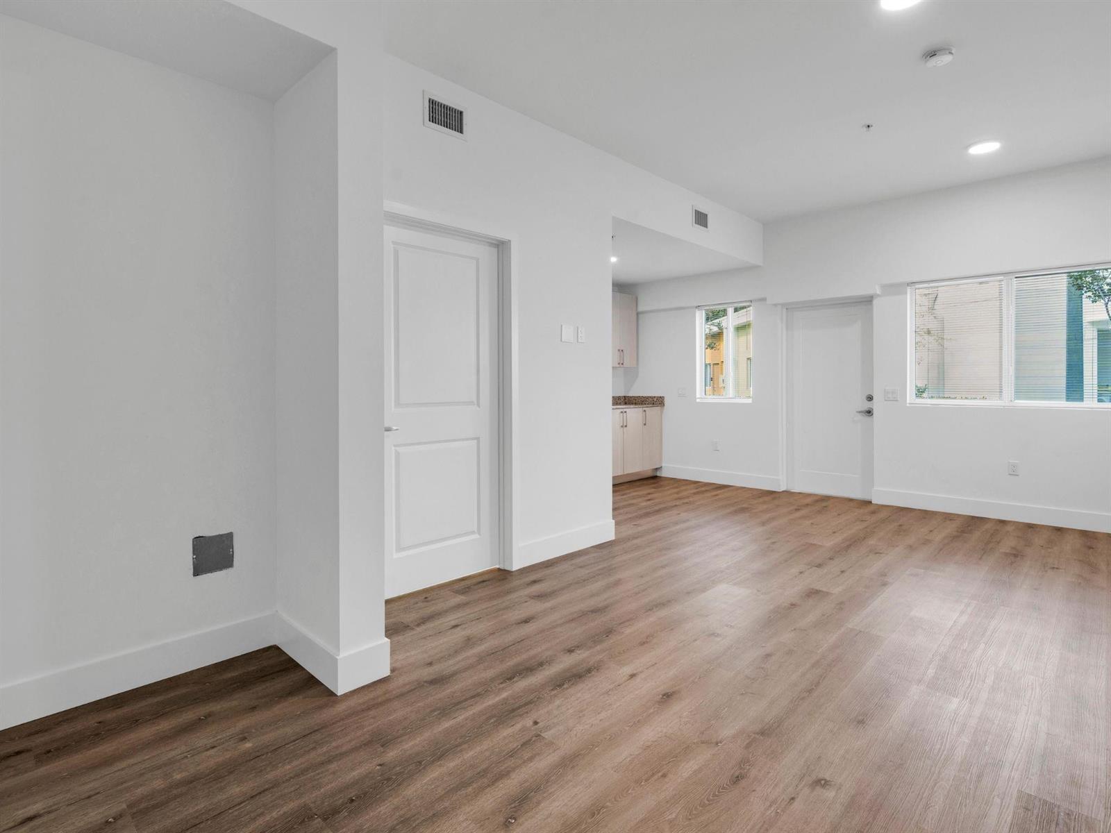 An empty room with white walls, wood-look flooring, a closed door, and a small kitchen area — 2130 NW 37th Street, Miami FL 33142, MIAMI, FL