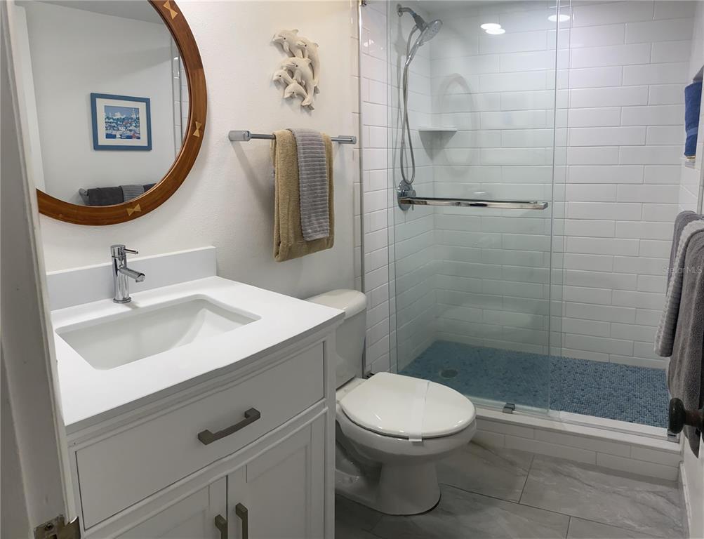A bathroom features a white vanity, a round mirror, a toilet, and a glass-enclosed shower — 1571 S ATLANTIC AVENUE UNIT 202, NEW SMYRNA BEACH, FL