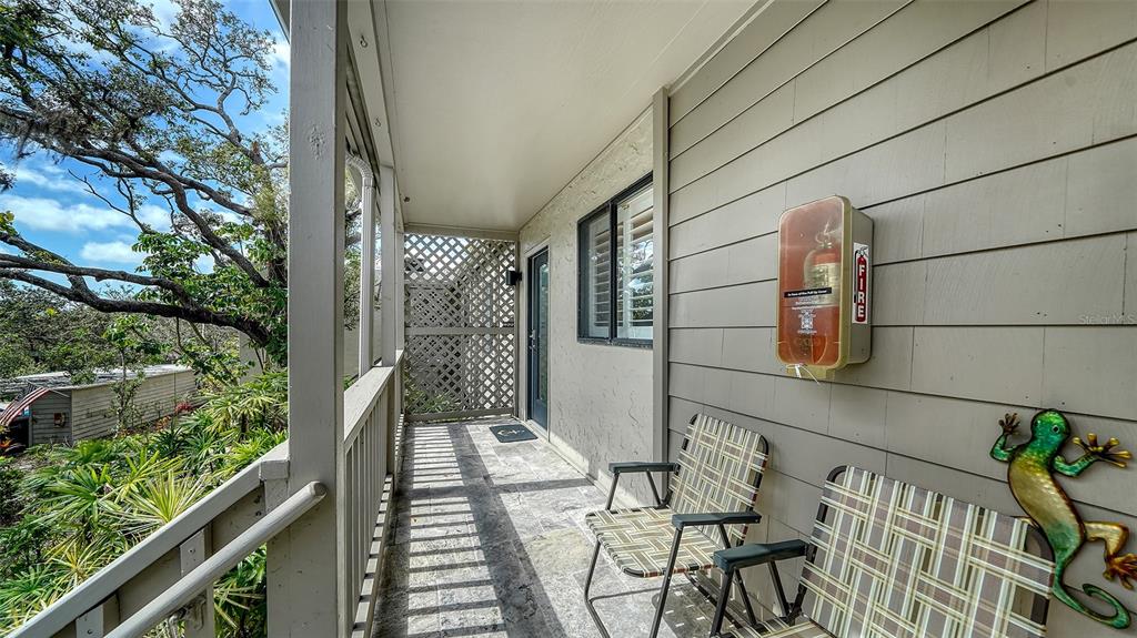 A covered outdoor balcony features a fire extinguisher, two chairs, and a decorative metal lizard — 1601 PELICAN POINT DRIVE UNIT HA212, SARASOTA, FL