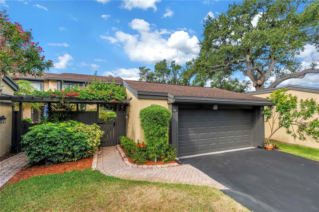 A tan house exterior features a dark gray garage door, a brick walkway, and landscaping — 13300 INDIAN ROCKS ROAD UNIT 103, LARGO, FL