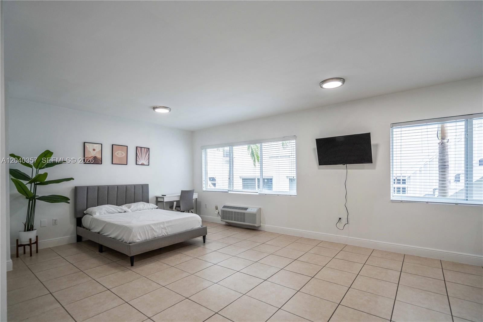 A bedroom features a gray upholstered bed, tile flooring, a wall-mounted television, and two windows — 6971 Carlyle Ave # 107, Miami Beach FL 33141, MIAMI BEACH, FL