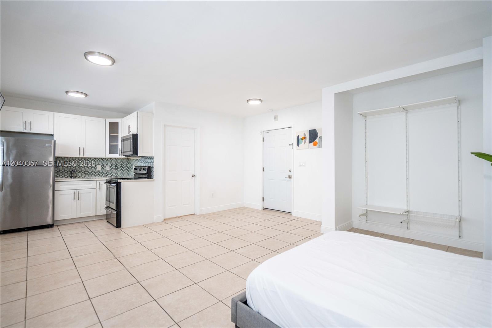 A studio apartment features a kitchen with white cabinets, tile flooring, and a bed area — 6971 Carlyle Ave # 107, Miami Beach FL 33141, MIAMI BEACH, FL